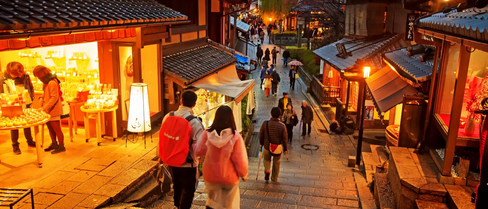Two people walk down stone steps in a bustling street, lined with traditional shops and warm lighting during dusk.