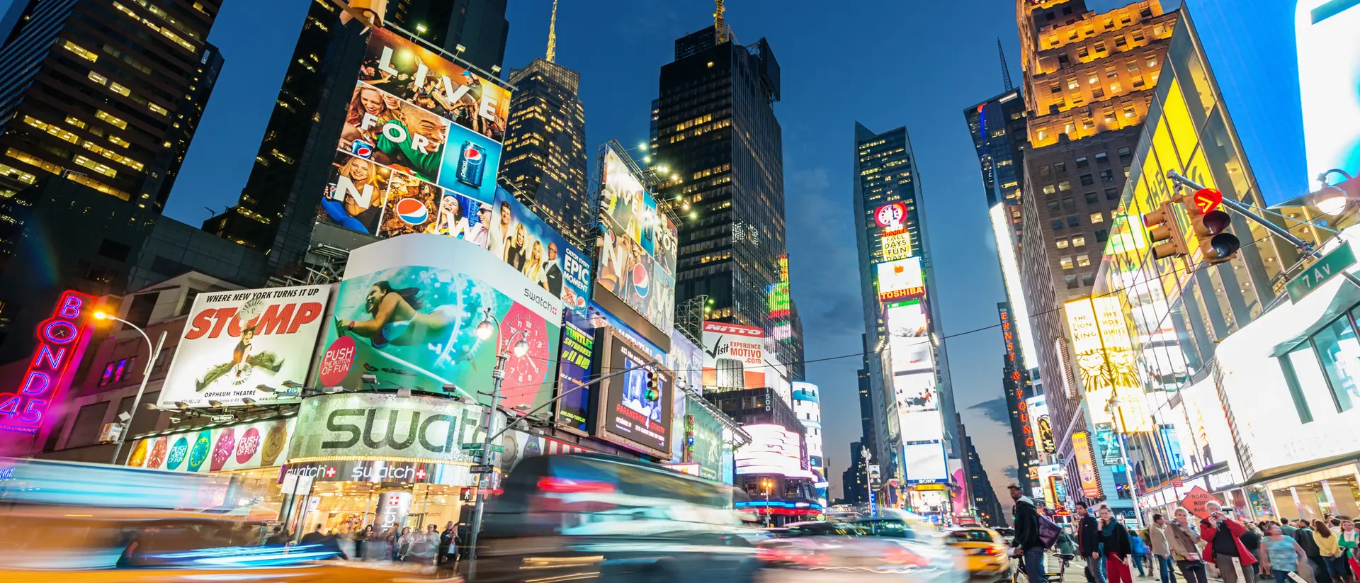 Brightly lit Times Square at dusk, with bustling traffic, pedestrians, and iconic billboards.