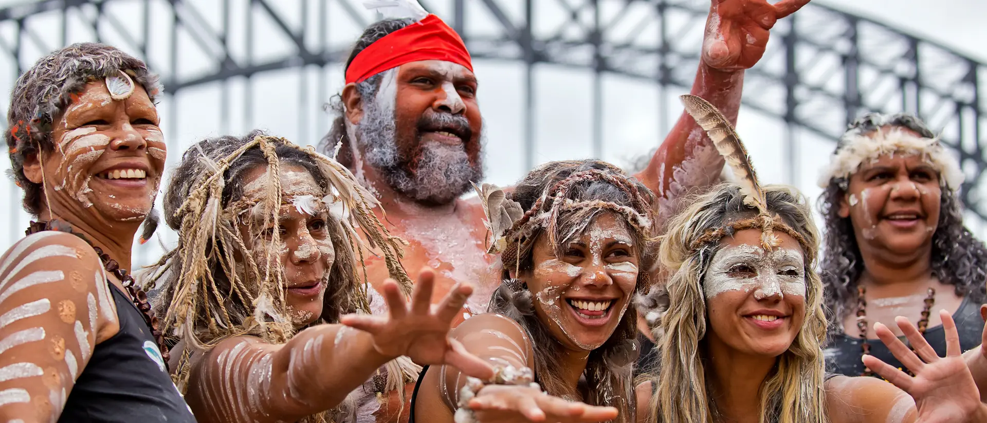 A group of six Indigenous people celebrating with traditional attire and body paint in front of a bridge.