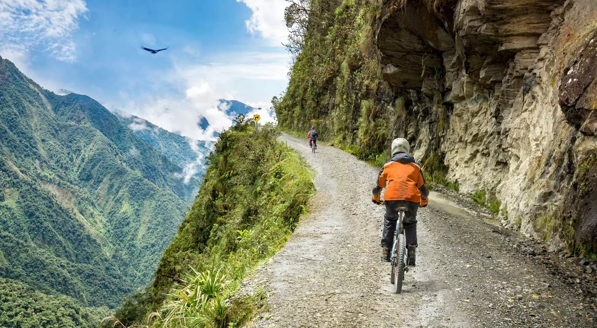 Bike tourists ride on North Yungas road, known as "Death Road" in Bolivia. In the background sky circles a condor over the scene.