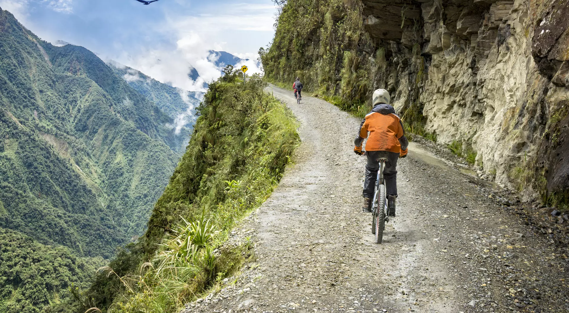 Two cyclists ride along a steep mountain path with lush greenery and a cloudy sky in the background.