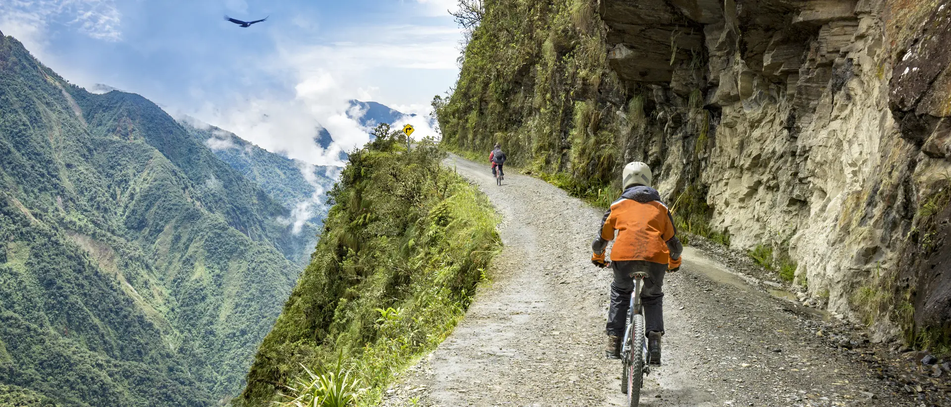 Two cyclists ride along a steep mountain path with lush greenery and a cloudy sky in the background.