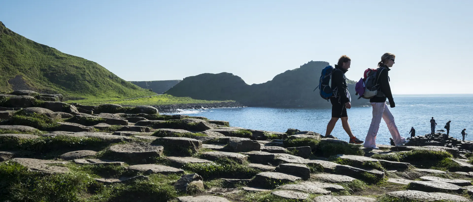 Two hikers walk along a rocky shoreline with green hills and water in the background on a sunny day.
