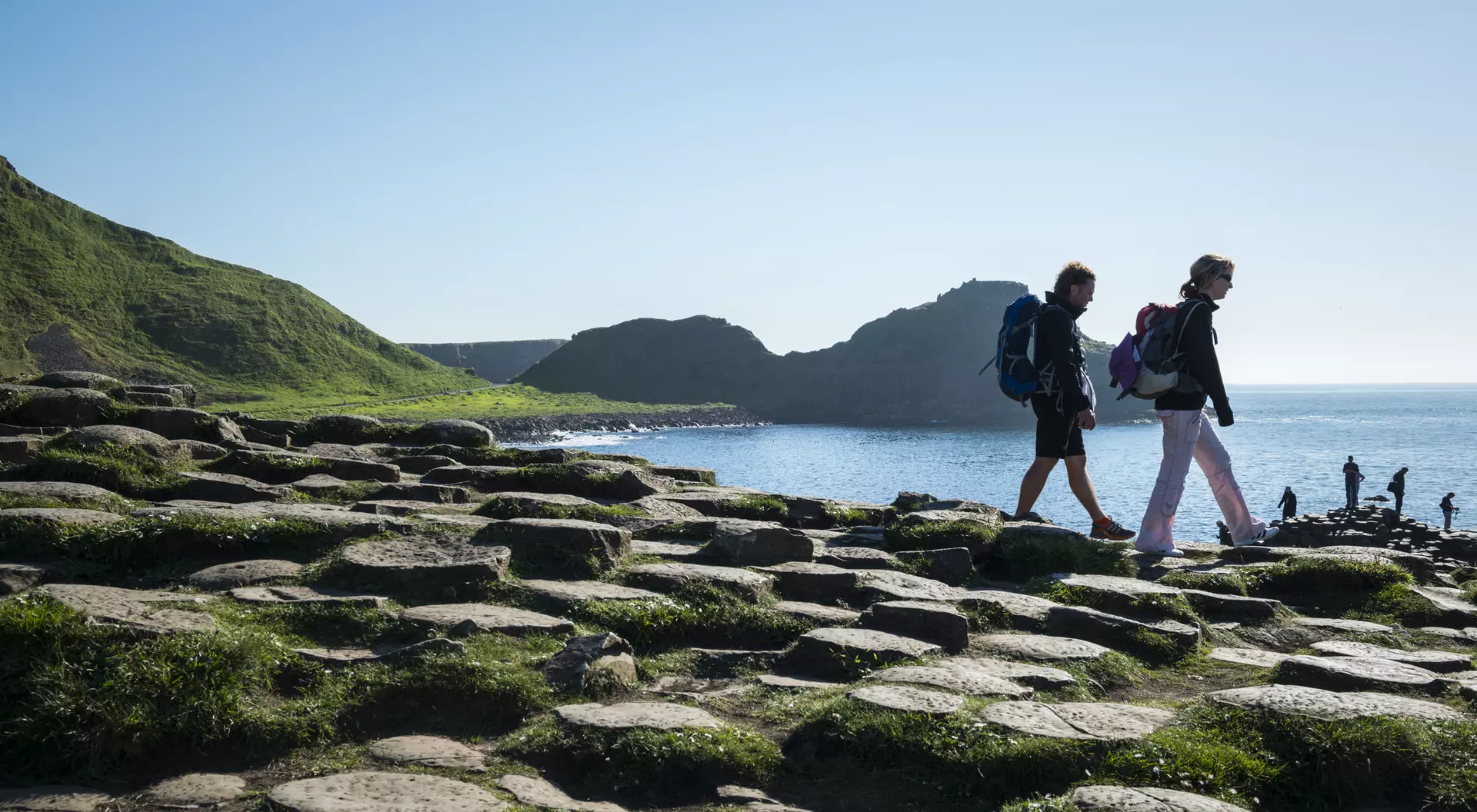 Two hikers walk along a rocky shoreline with green hills and water in the background on a sunny day.