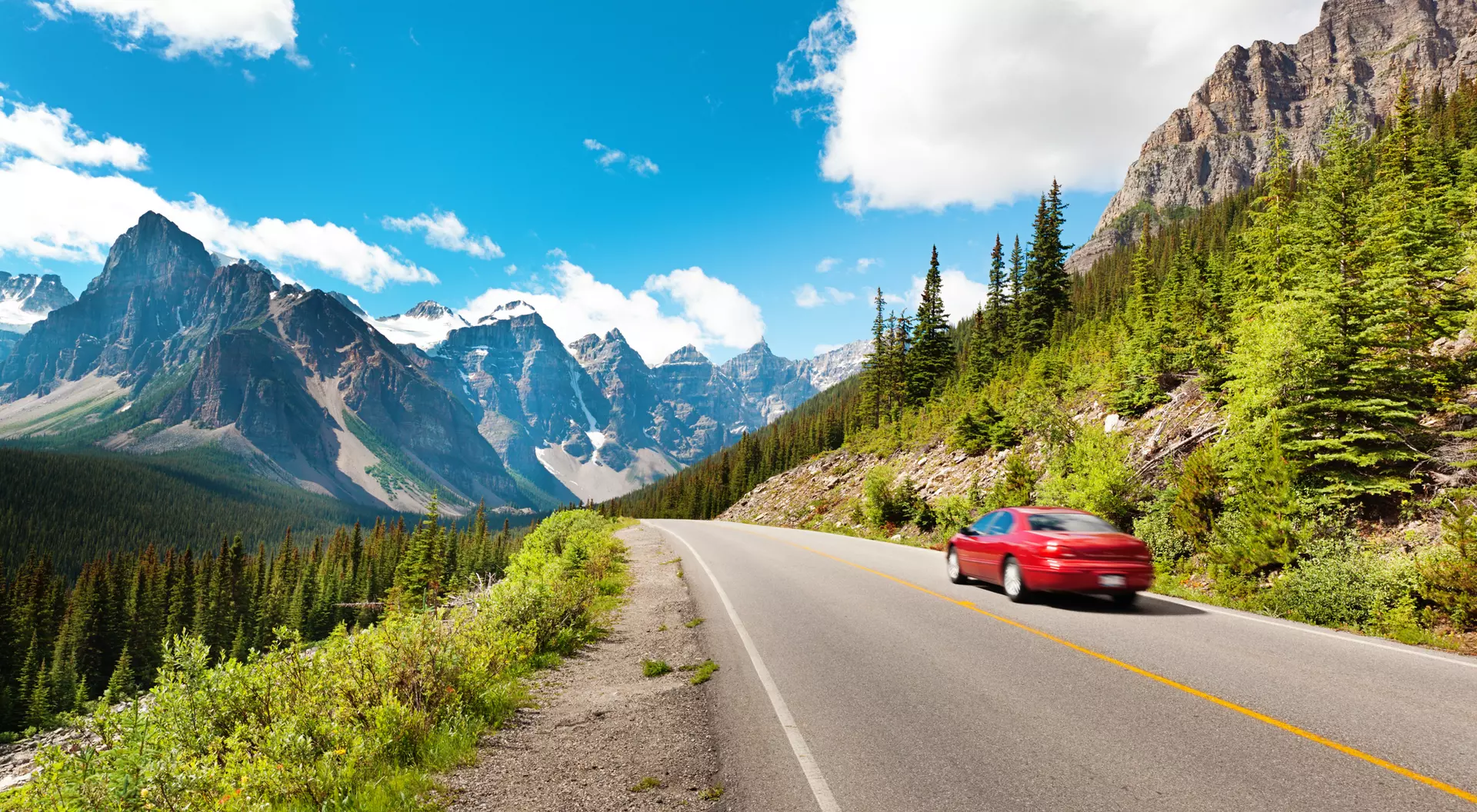 A red car drives along a winding road with majestic mountains and green trees under a bright blue sky.