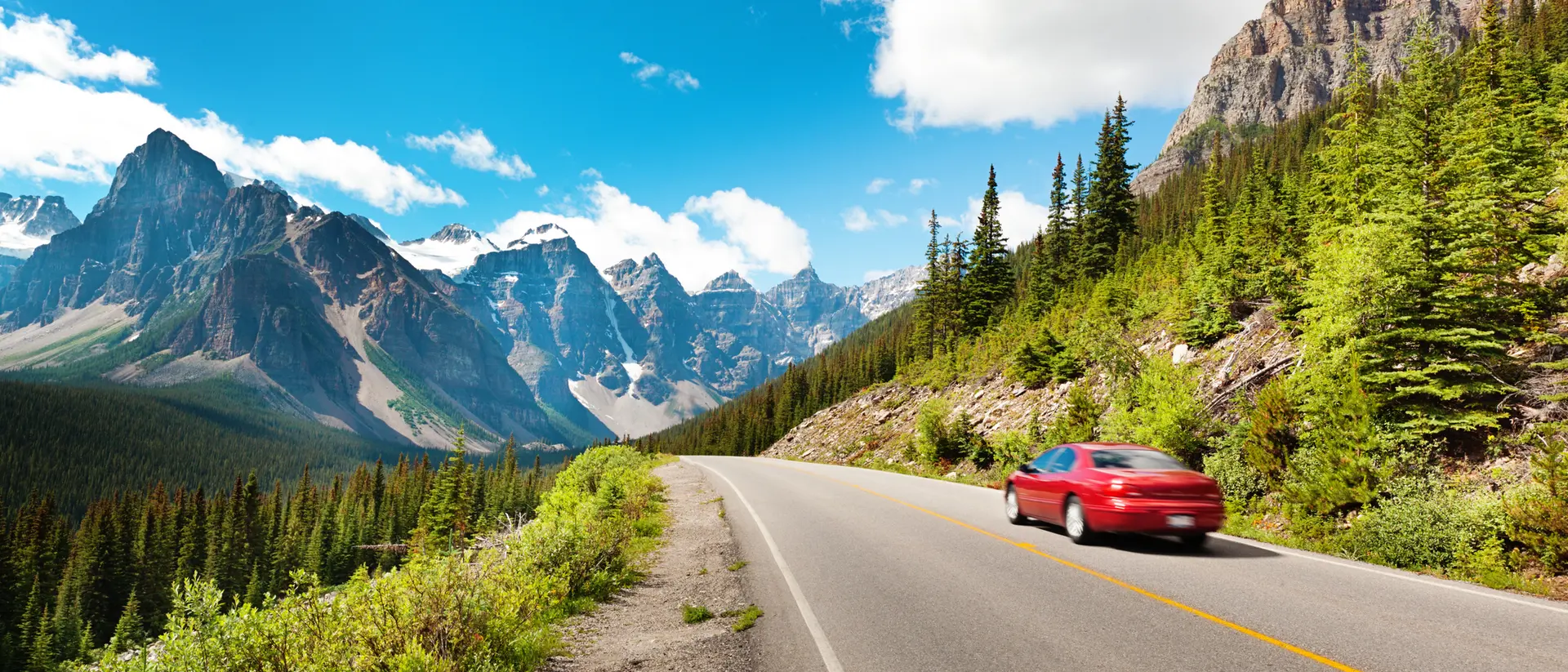 A red car drives along a winding road with majestic mountains and green trees under a bright blue sky.