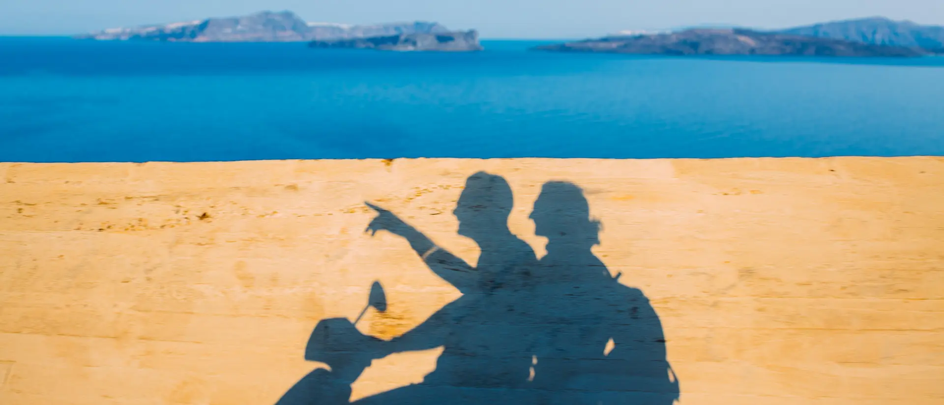 Shadow of two people on a motorcycle, overlooking a blue sea and distant islands on a sunny day.