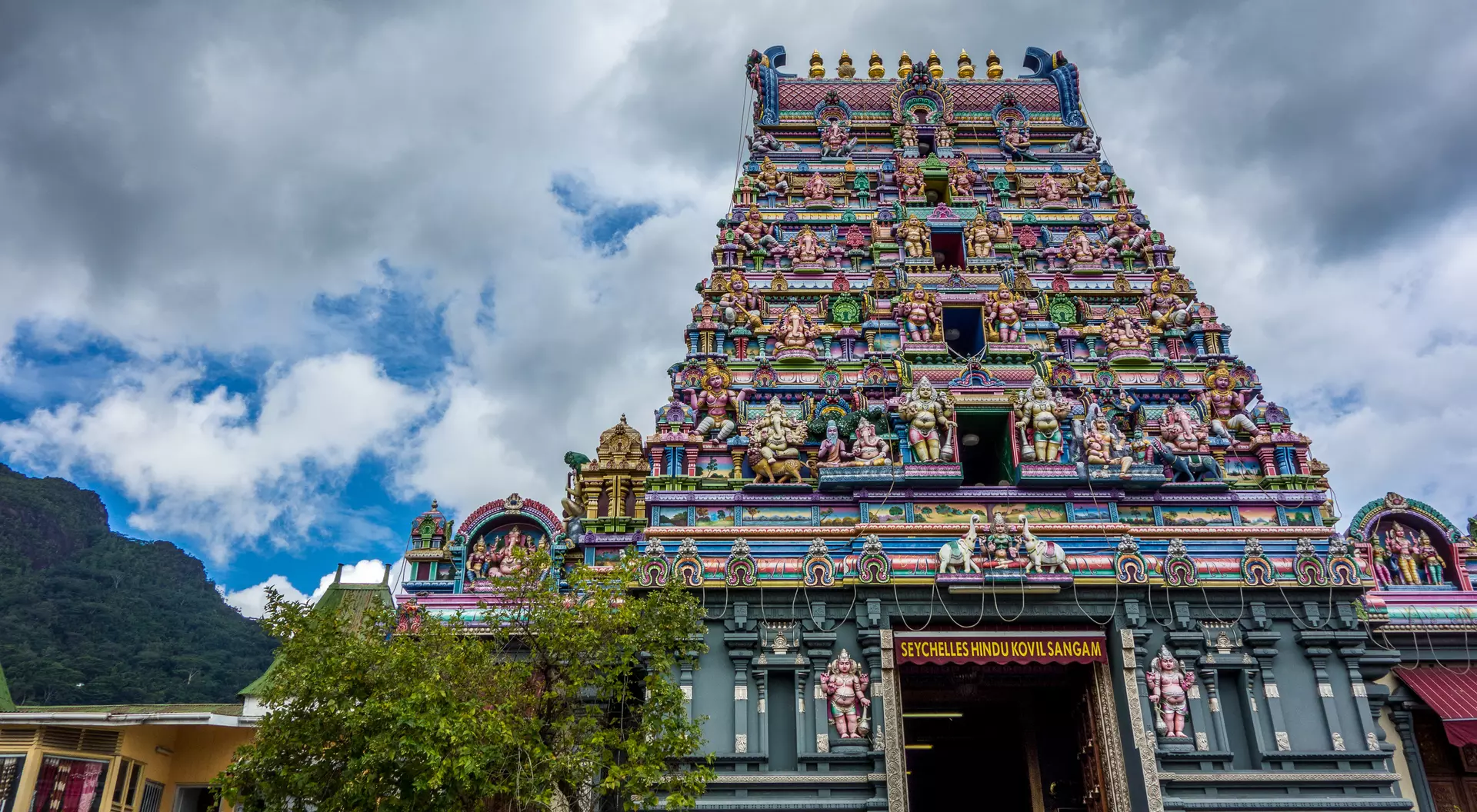 Colorful temple tower with intricate sculptures, set against a cloudy sky and lush greenery below.