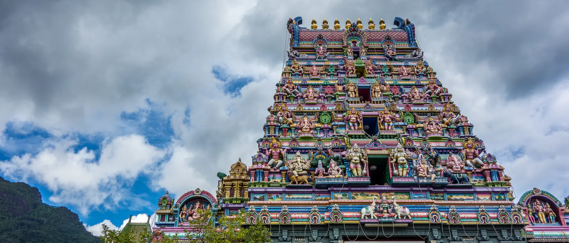 Colorful temple tower with intricate sculptures, set against a cloudy sky and lush greenery below.