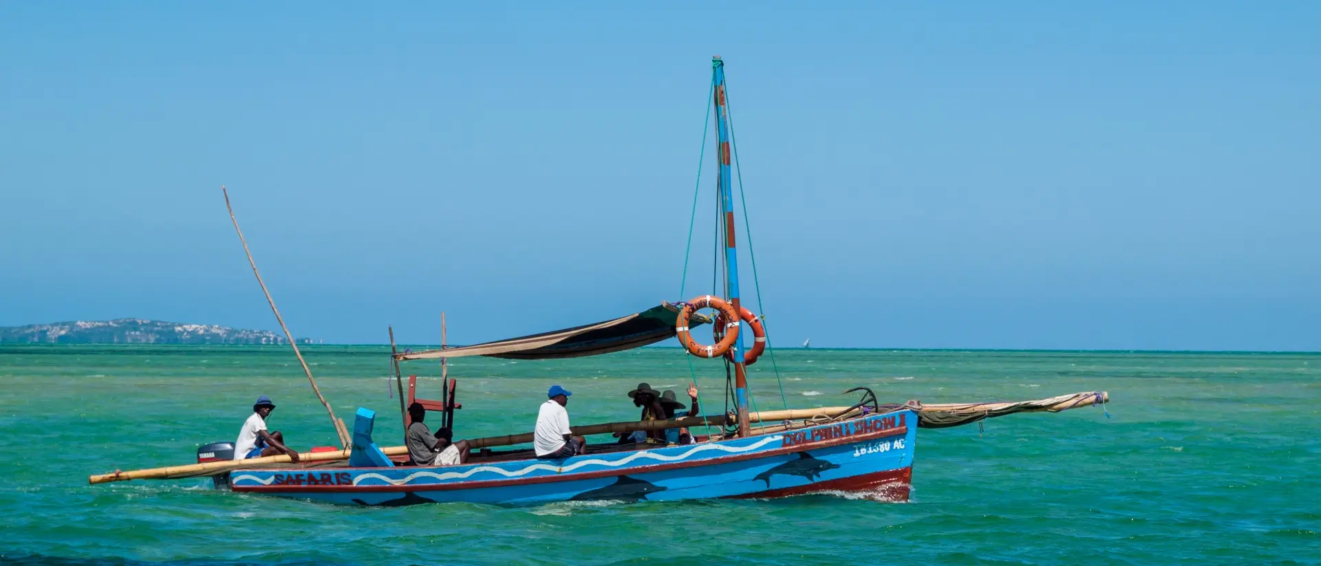 A colorful fishing boat with several people on the water, surrounded by a bright blue sky and clear green sea.