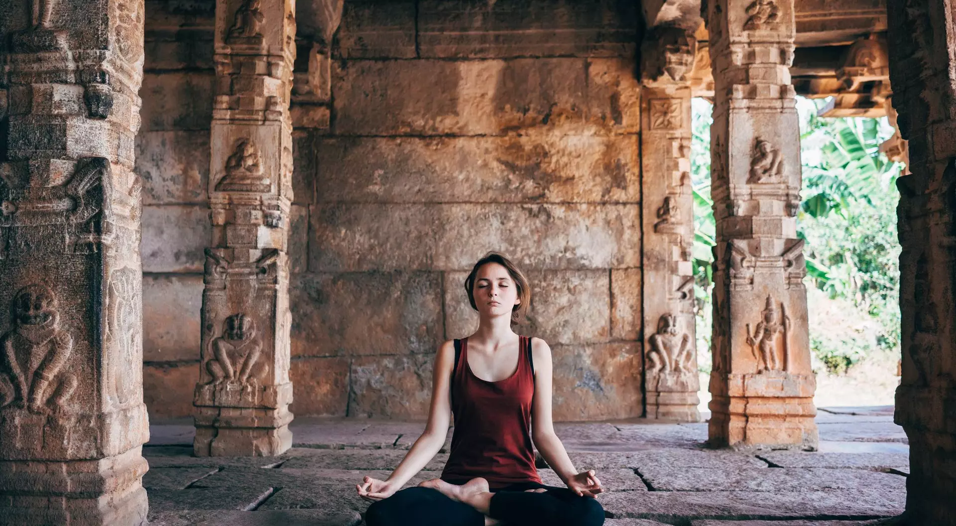 A woman meditating in a serene temple with stone pillars and lush greenery in the background.