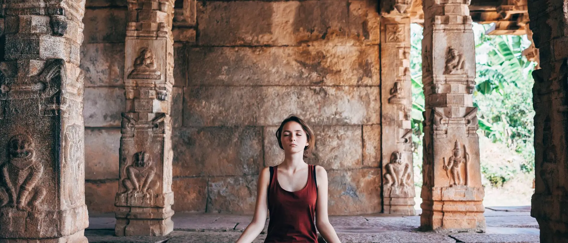 A woman meditating in a serene temple with stone pillars and lush greenery in the background.