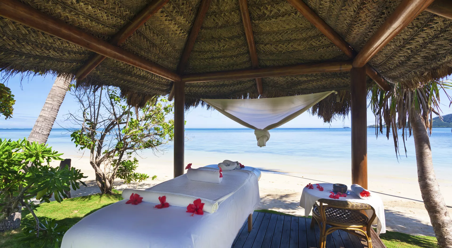 Spa treatment area with massage tables under a thatched roof, overlooking a serene beach and clear blue water.