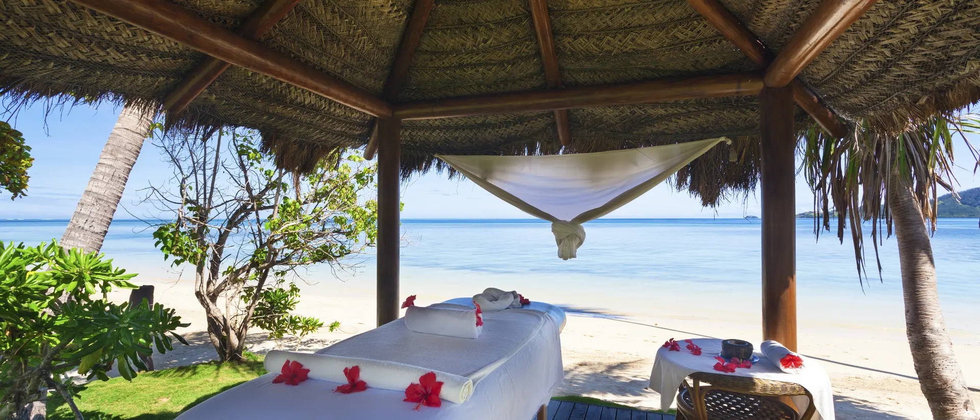 Spa treatment area with massage tables under a thatched roof, overlooking a serene beach and clear blue water.