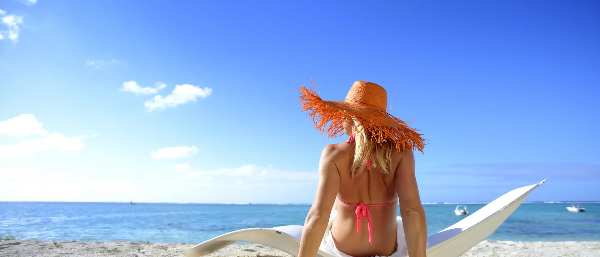 A person sits on the beach, wearing a large straw hat and a pink bikini, facing the ocean under a clear blue sky.