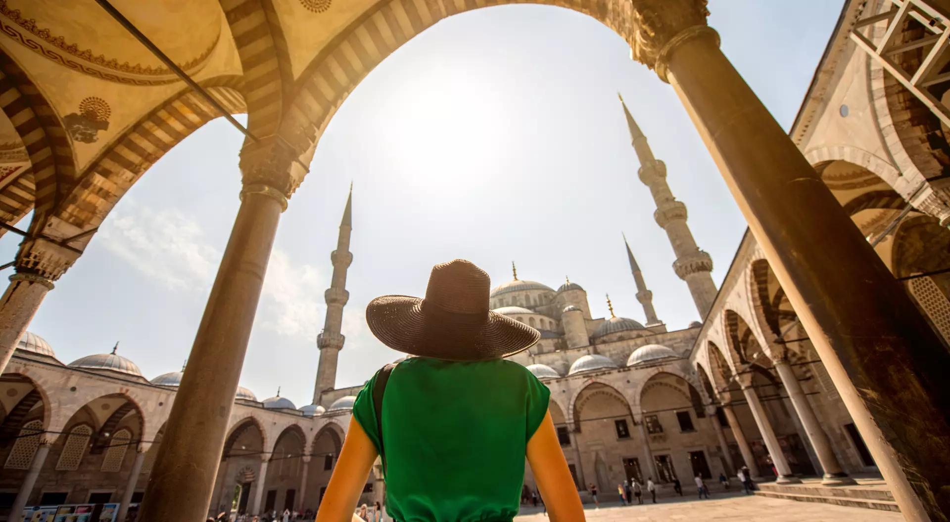 Person in a green dress and wide-brimmed hat stands under arches, overlooking a mosque with tall minarets and a bright sky.