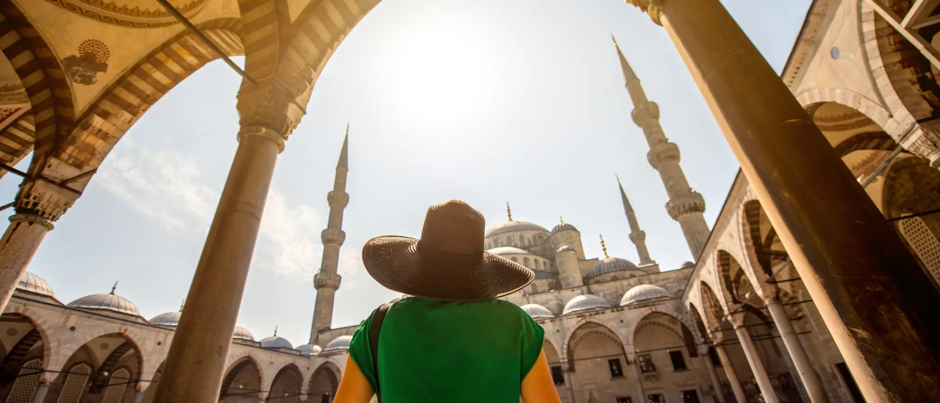 Person in a green dress and wide-brimmed hat stands under arches, overlooking a mosque with tall minarets and a bright sky.