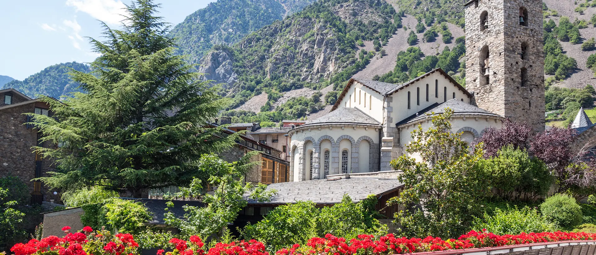 A scenic view of a stone church and tower surrounded by greenery and flowers, set against a mountain backdrop.