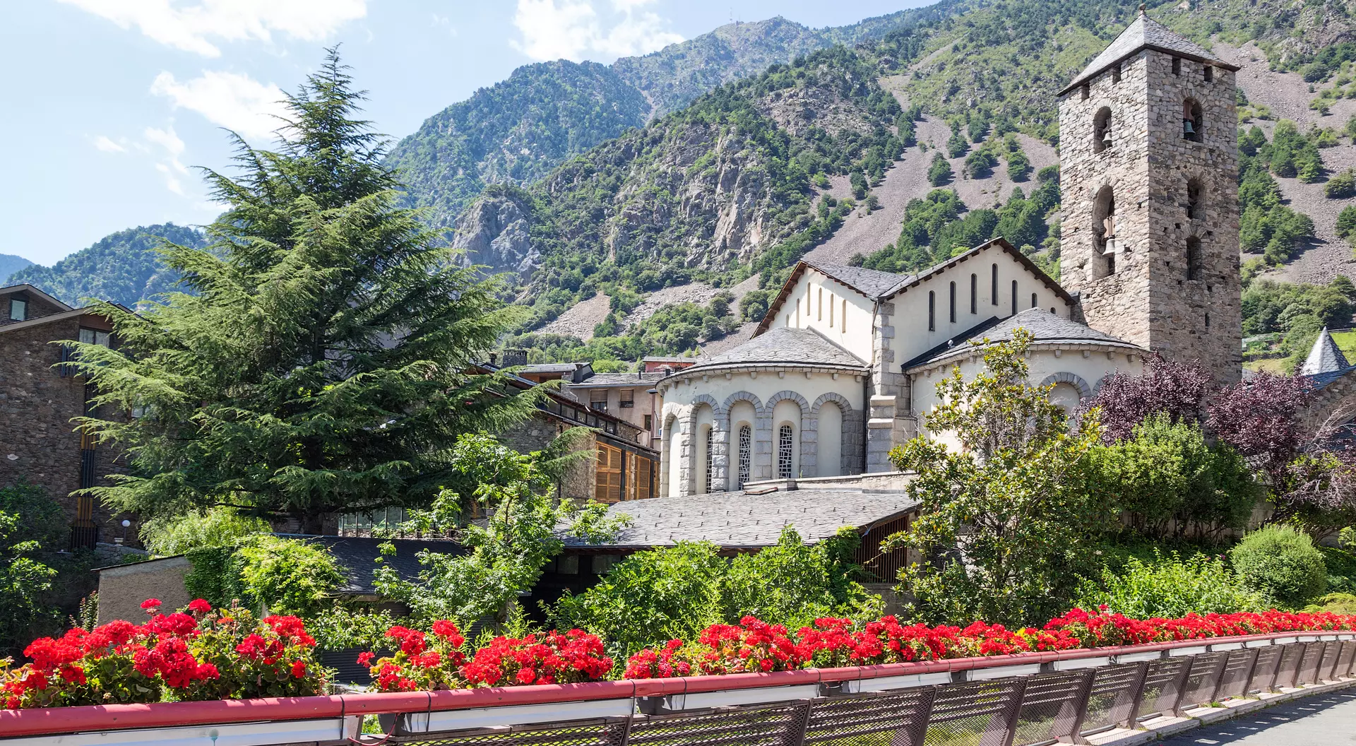 A scenic view of a stone church and tower surrounded by greenery and flowers, set against a mountain backdrop.