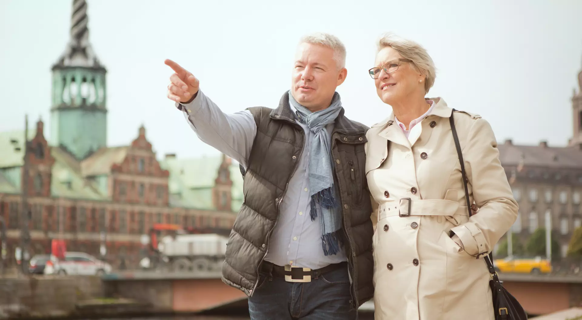 A smiling couple stands by a river, with the man pointing while the woman looks at him, featuring historic buildings in the background.