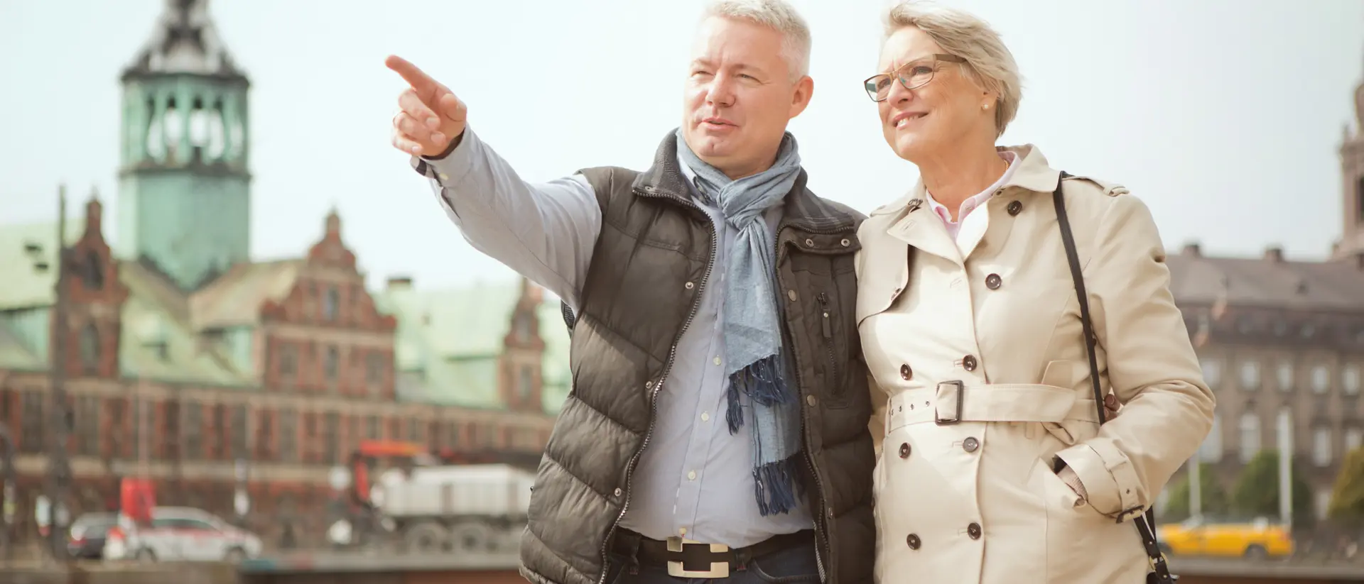 A smiling couple stands by a river, with the man pointing while the woman looks at him, featuring historic buildings in the background.