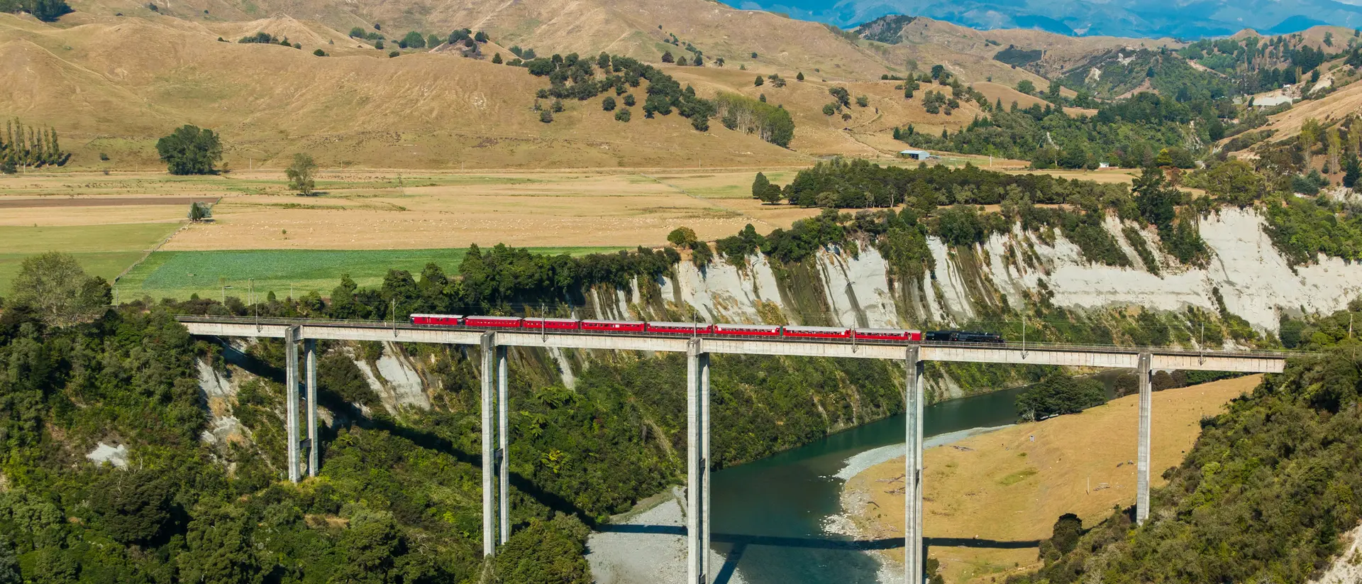 A red train crosses a tall bridge over a river, surrounded by green forests and rolling hills under a blue sky.
