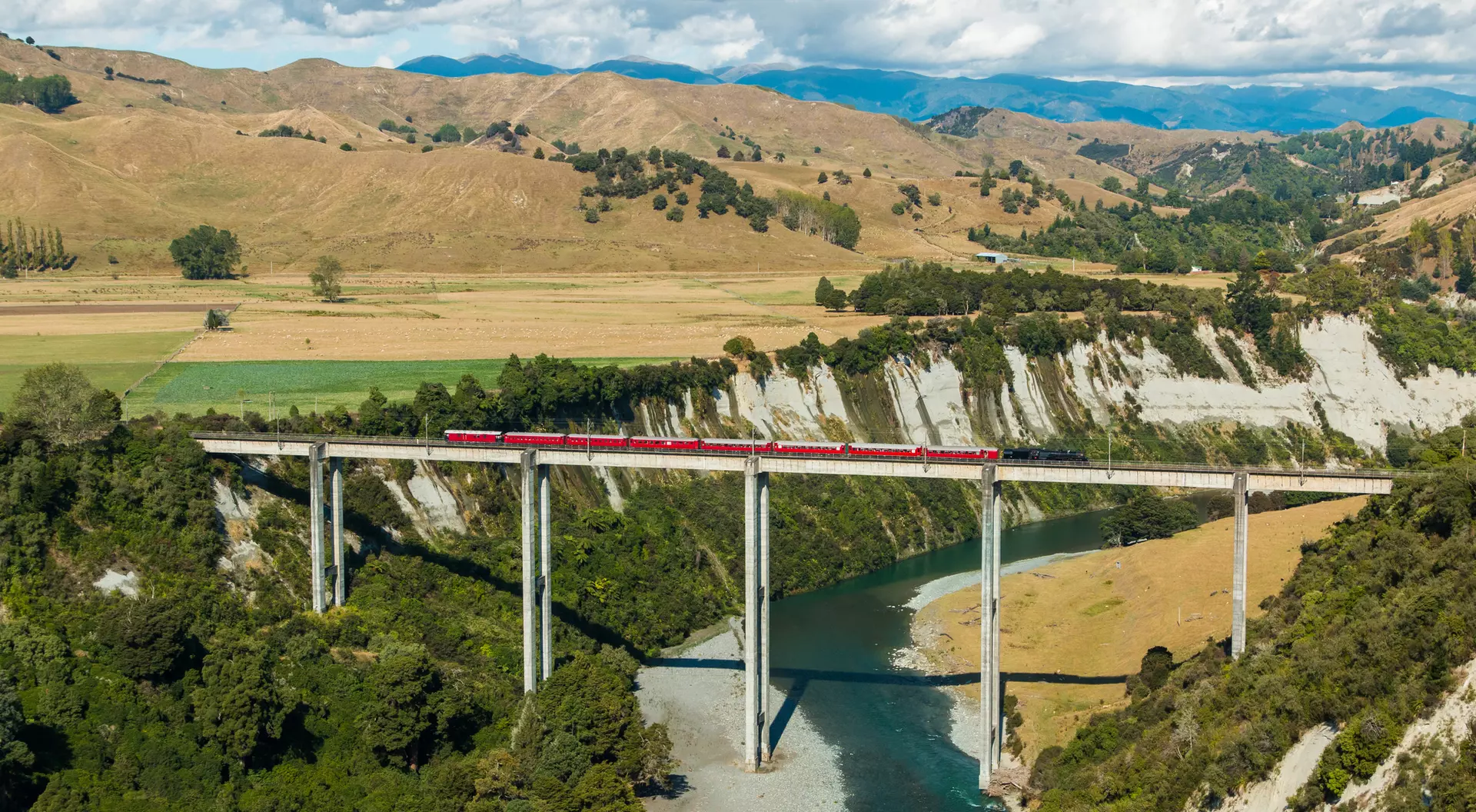 A red train crosses a tall bridge over a river, surrounded by green forests and rolling hills under a blue sky.
