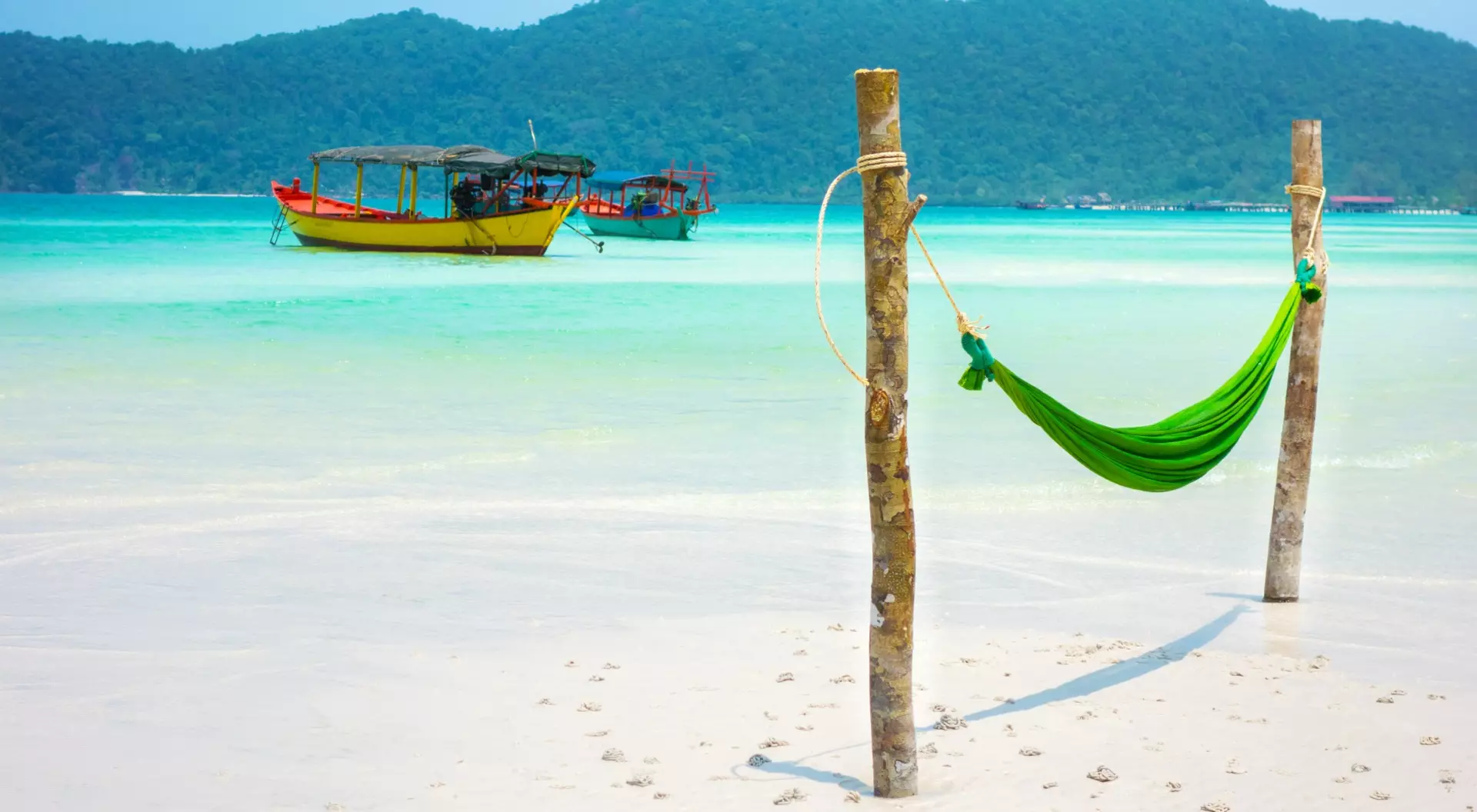 A green hammock between two wooden posts on a sandy beach, with boats and hills in the background.