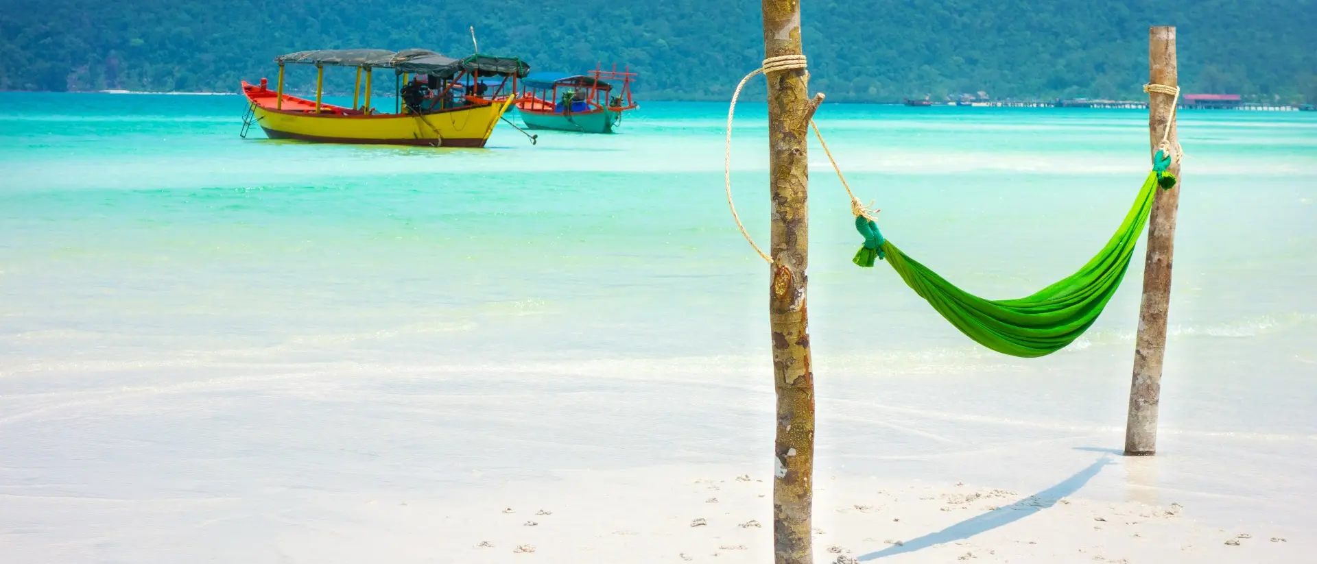 A green hammock between two wooden posts on a sandy beach, with boats and hills in the background.