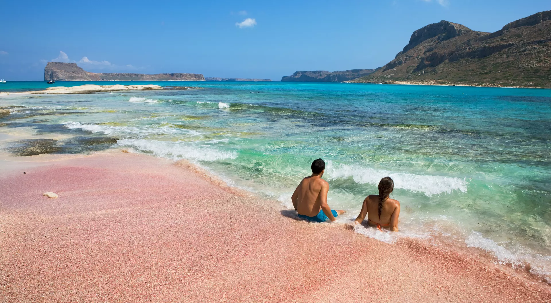 Two people sitting on a pink beach, gazing at turquoise waters and distant hills under a clear blue sky.