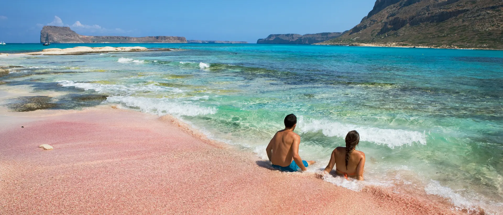 Two people sitting on a pink beach, gazing at turquoise waters and distant hills under a clear blue sky.