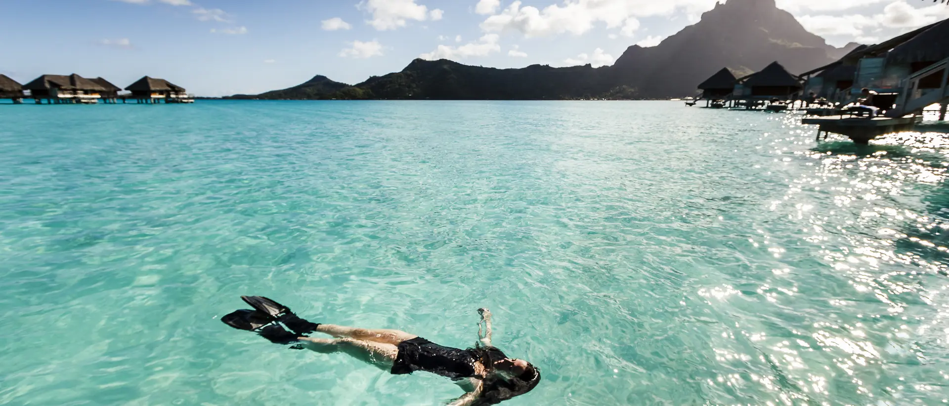 Person in snorkel gear floats peacefully in turquoise water, surrounded by mountains and overwater bungalows under a sunny sky.
