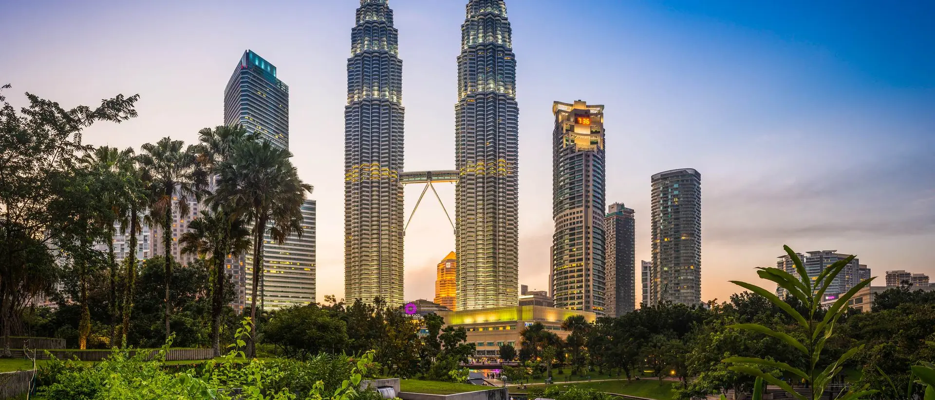 Petronas Towers rise above lush greenery at dusk, with city skyline in the background, showcasing modern architecture.