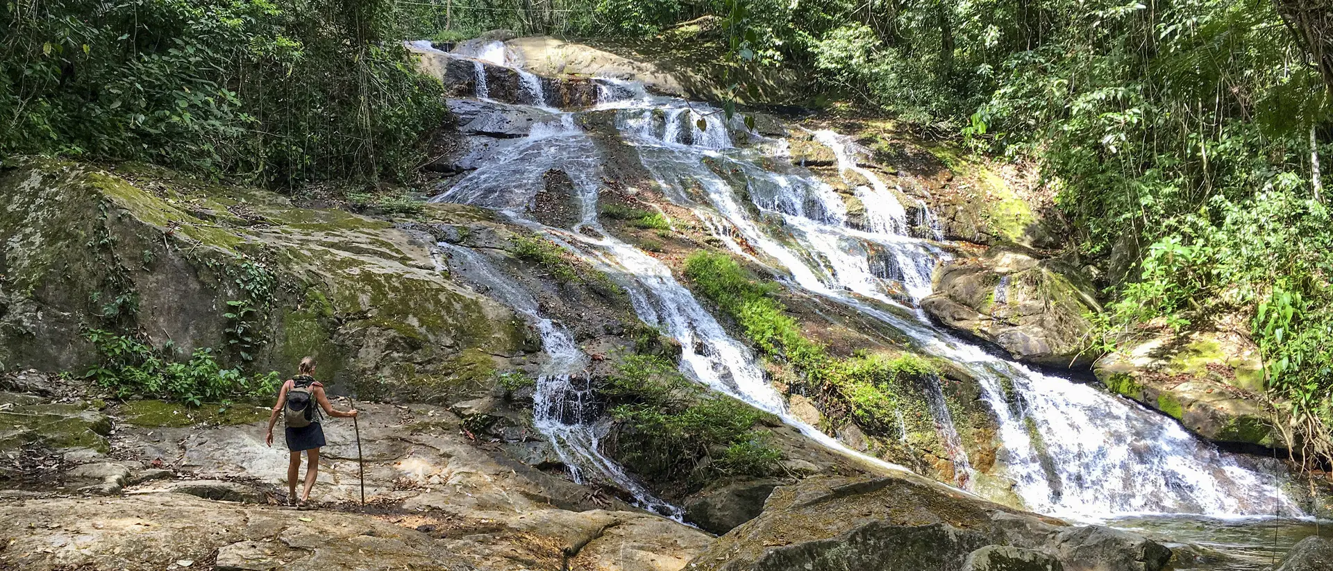 A hiker stands on rocky terrain near a cascading waterfall surrounded by lush greenery.