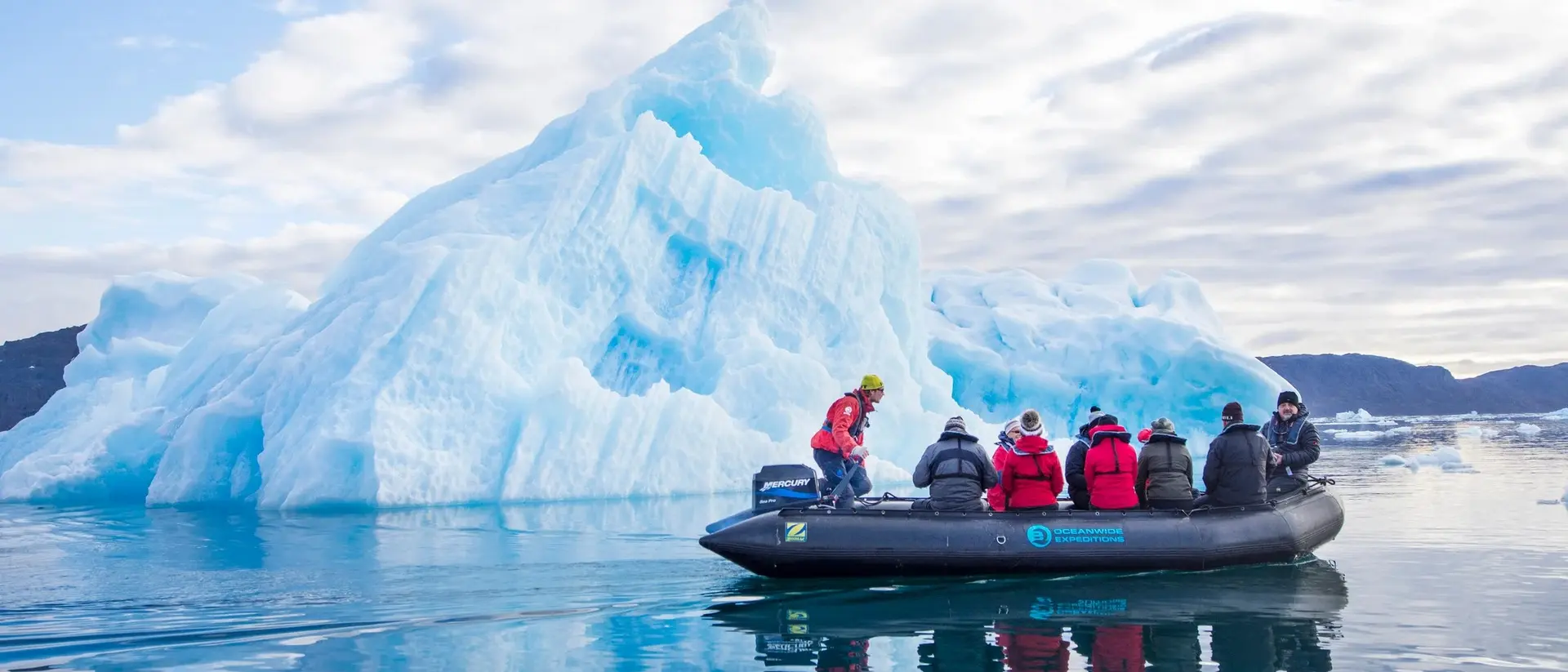 A group of people in a boat near a large blue iceberg on calm water under a cloudy sky.
