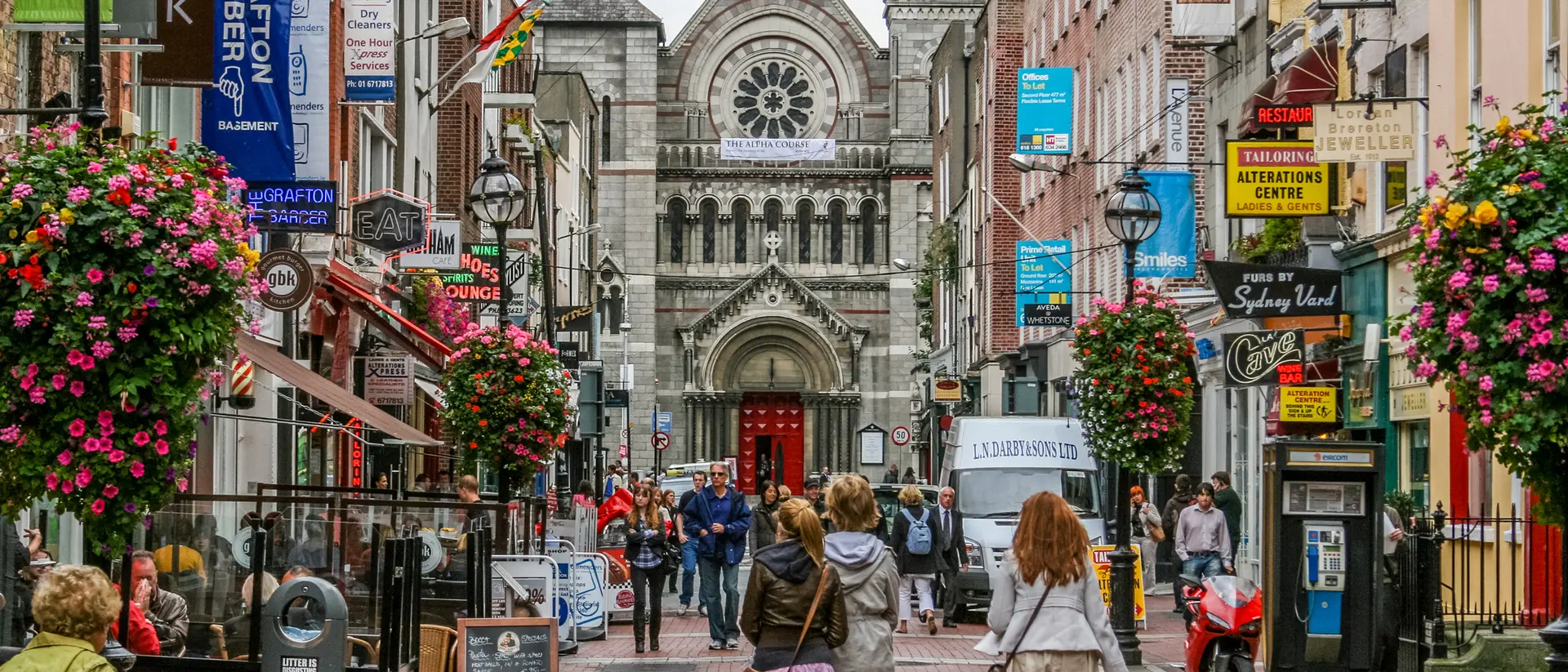 Bustling city street lined with shops and colorful flowers, leading to a historic church in the background.