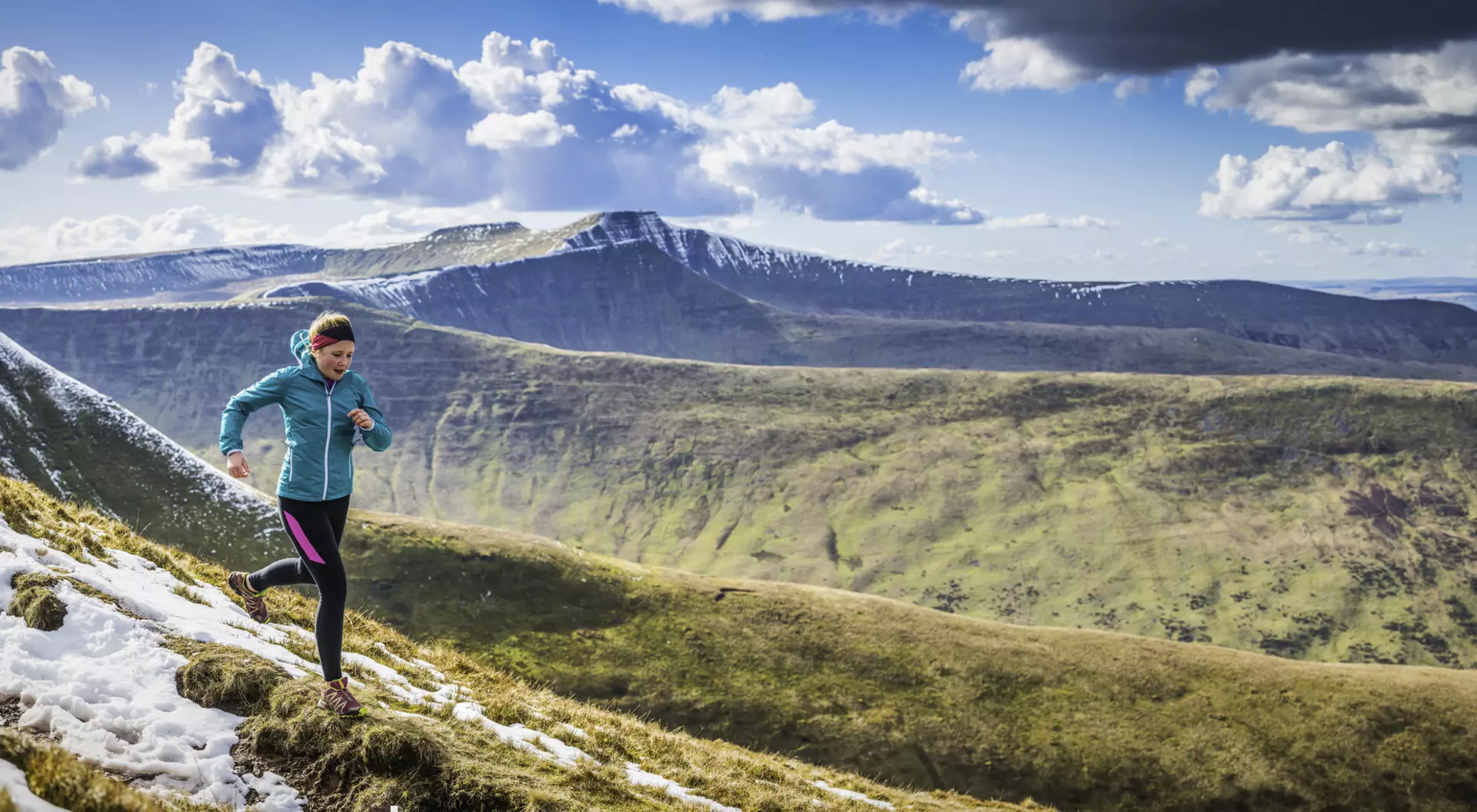 A person runs on a snowy mountain trail with scenic hills and clouds in the background.
