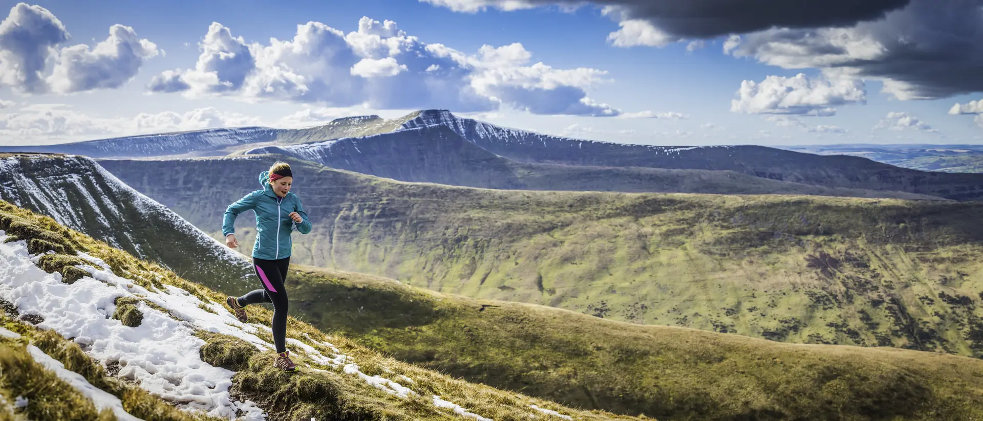 A person runs on a snowy mountain trail with scenic hills and clouds in the background.
