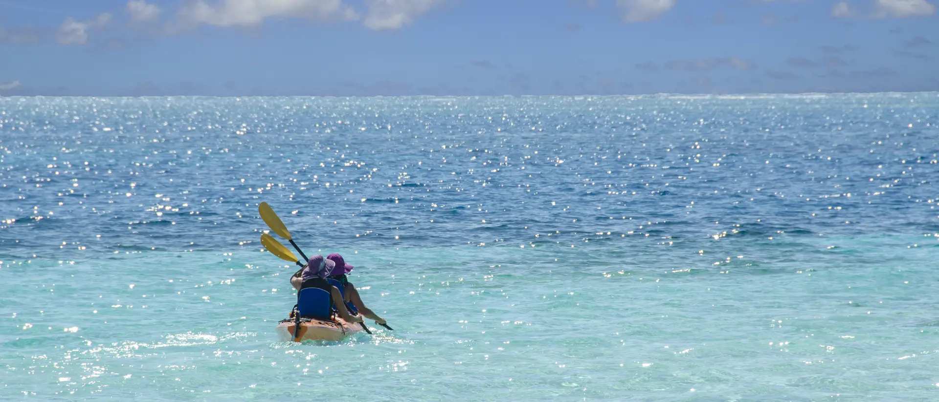 A person paddling a kayak in clear blue water under a bright sky with fluffy clouds.