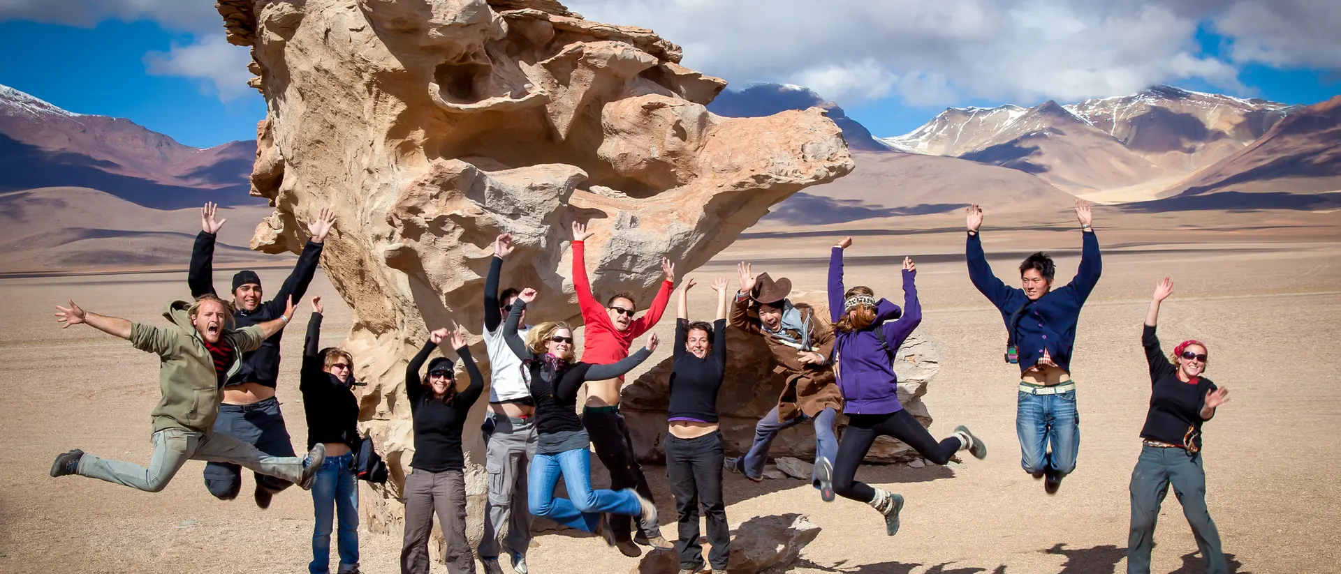 Group of people joyfully jumping in front of a large rock formation in a desert landscape with mountains in the background.