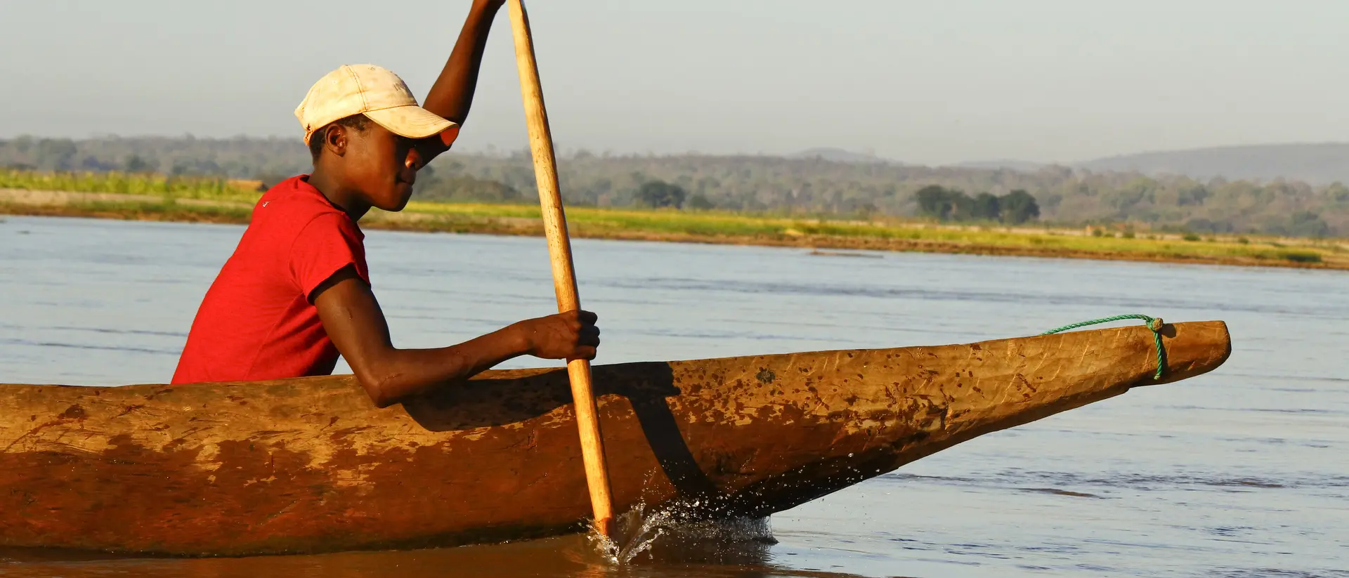 A person in a red shirt paddles a wooden canoe on a calm river during daylight.