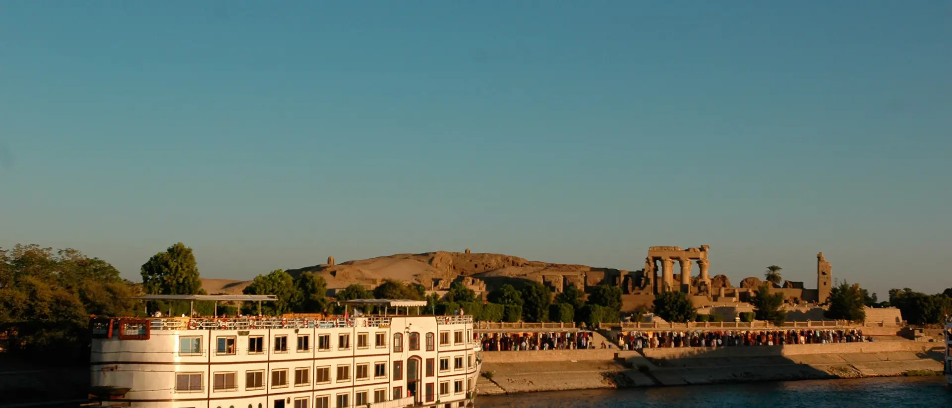 A riverboat anchored near ancient ruins, with clear blue skies and people gathered along the shoreline in the background.