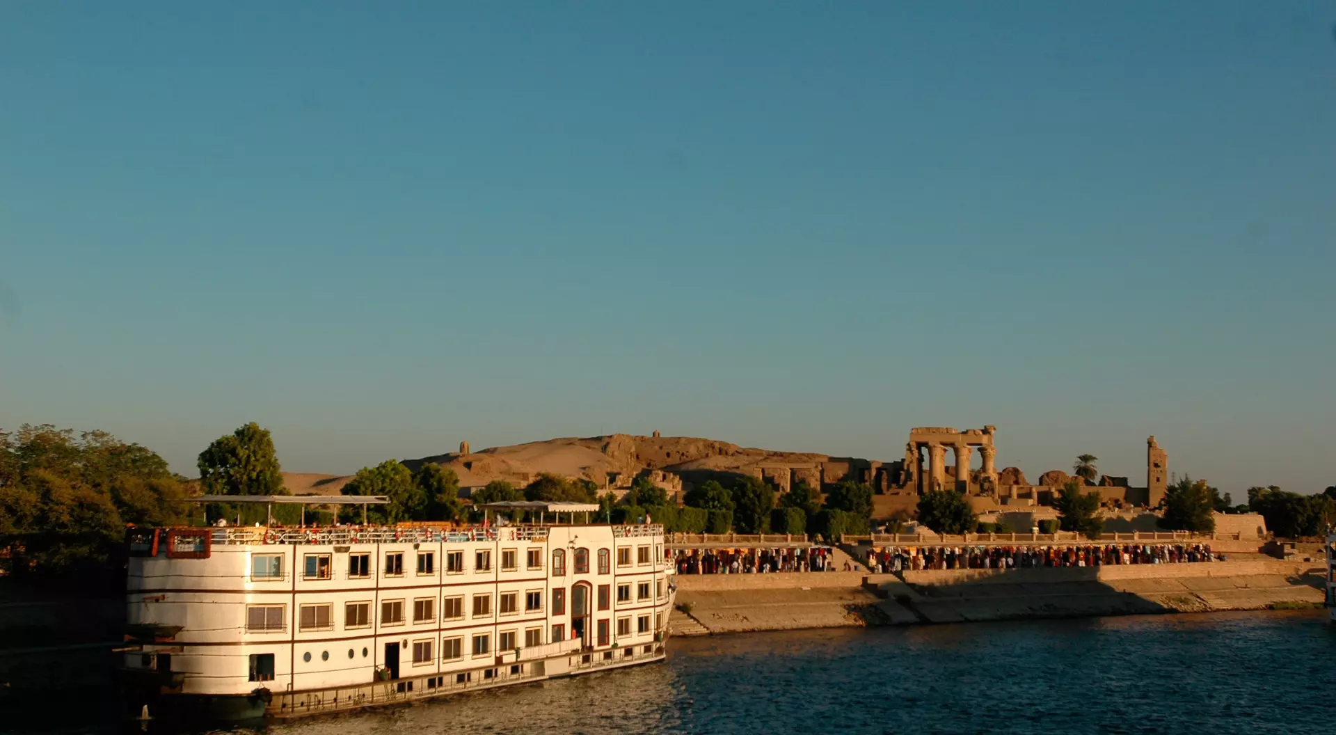 A riverboat anchored near ancient ruins, with clear blue skies and people gathered along the shoreline in the background.