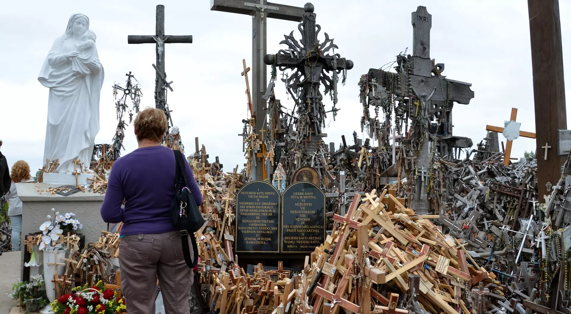 A woman stands before a shrine filled with wooden crosses and a statue, reflecting a memorial site.