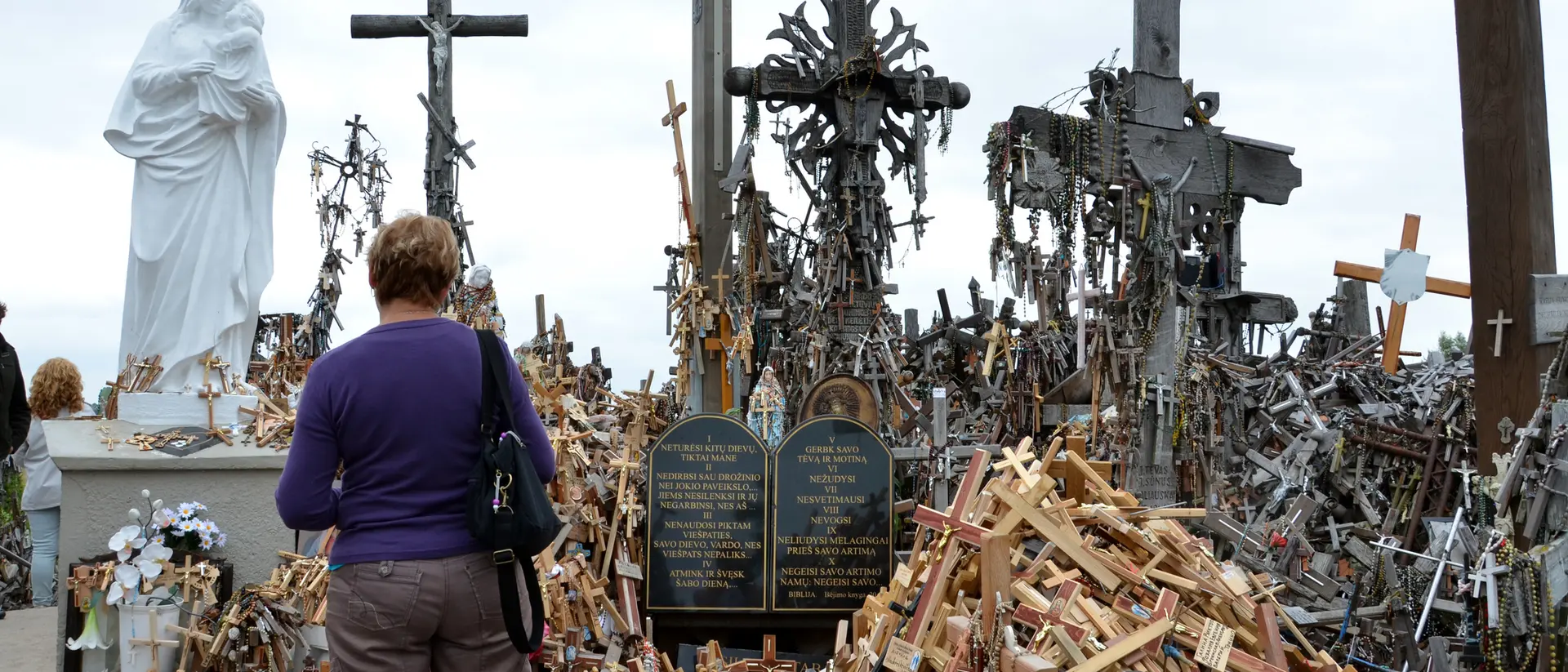 A woman stands before a shrine filled with wooden crosses and a statue, reflecting a memorial site.