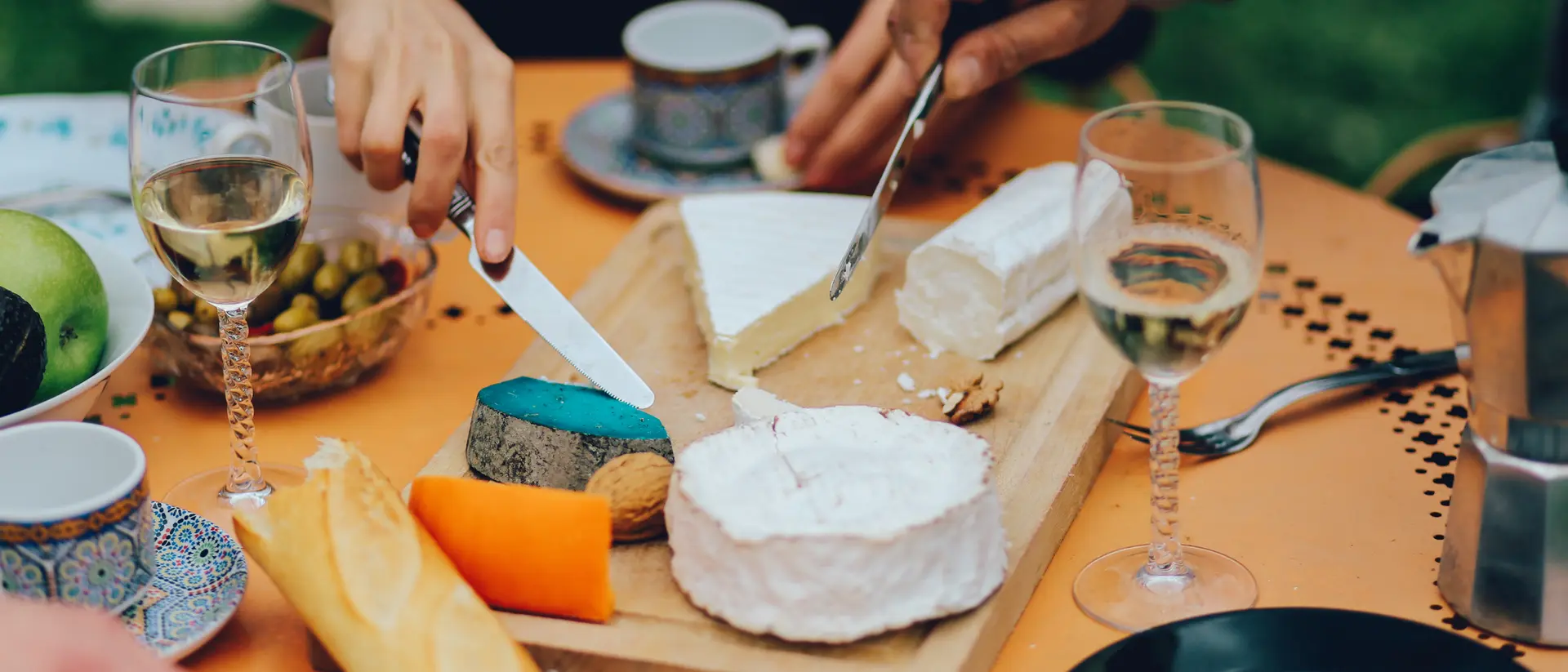 A close-up of a cheese platter with various cheeses, wine glasses, and fruits on a table. Hands are cutting cheese.