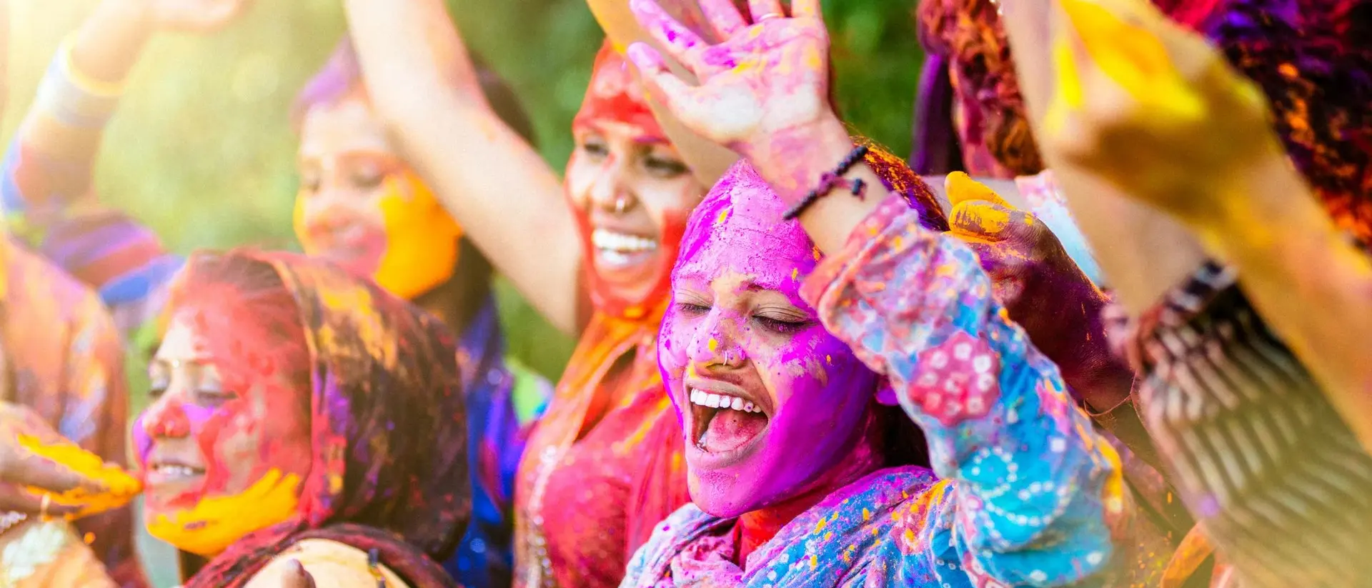 A group of joyful women celebrate with bright colors during a festival, smiling and raising their hands in excitement.