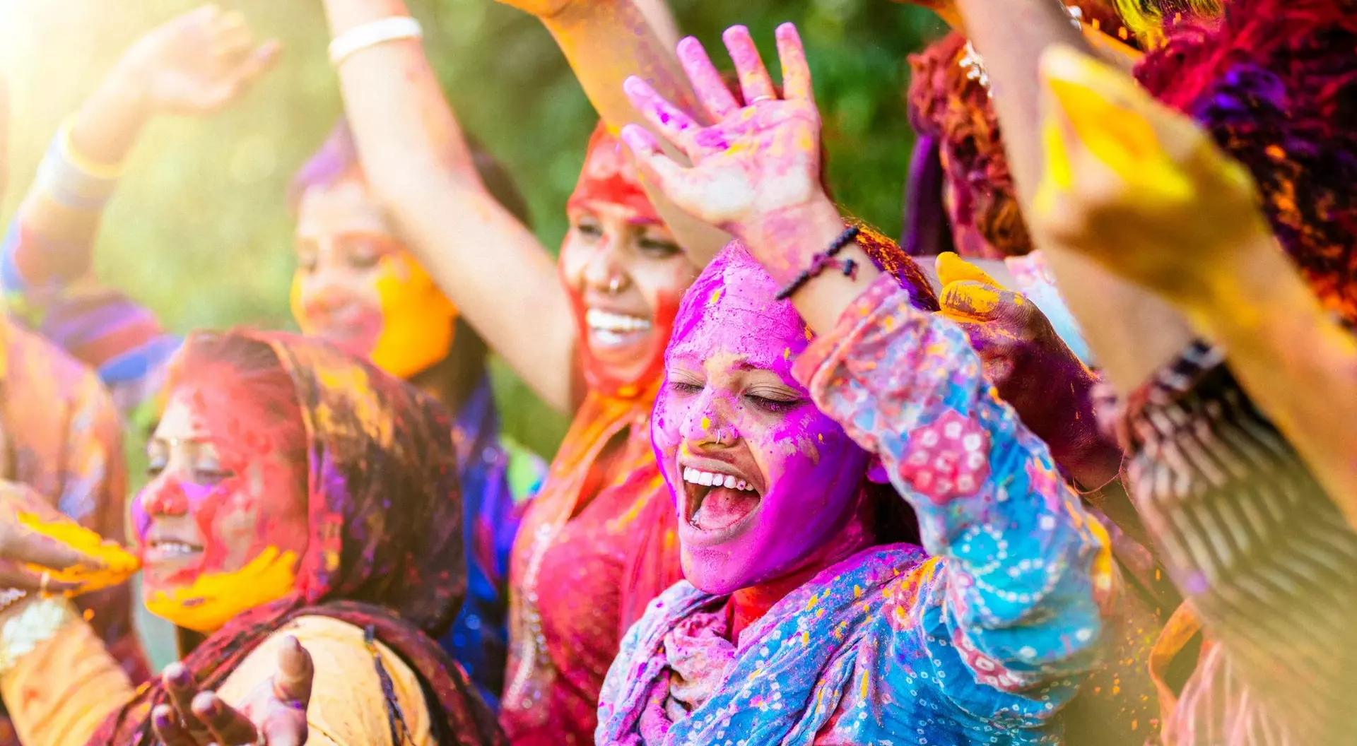 A group of joyful women celebrate with bright colors during a festival, smiling and raising their hands in excitement.