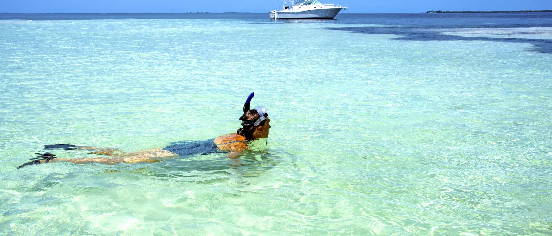 A person snorkels in clear turquoise water, with a boat in the background and a blue sky above.