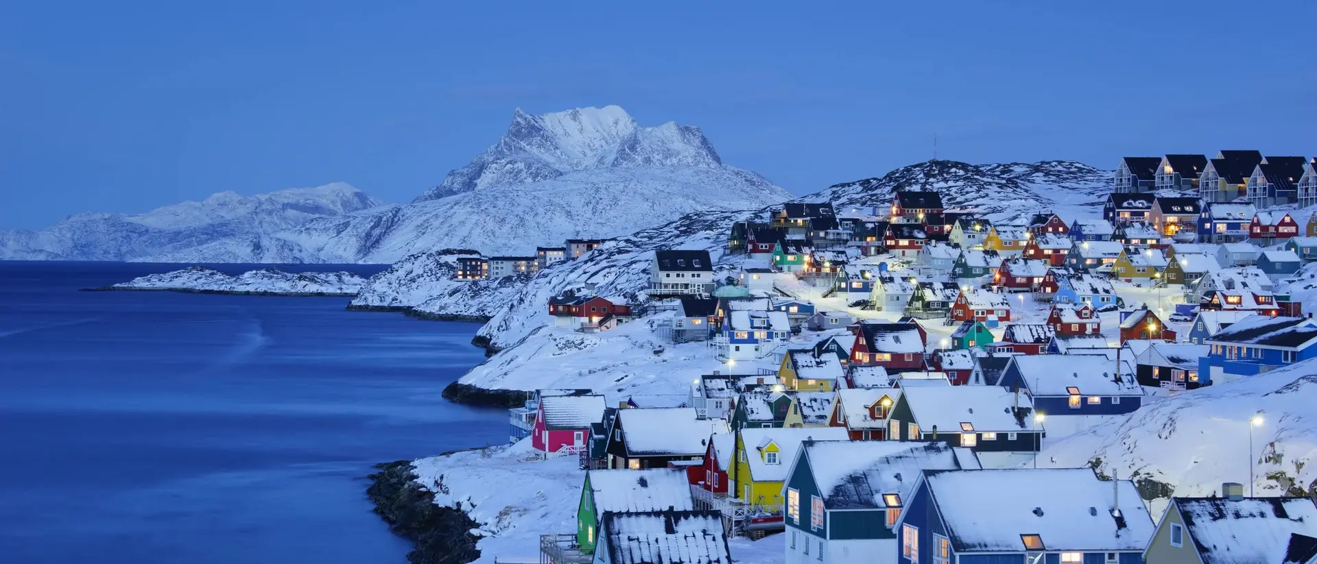 Colorful snowy houses line a coastal hill, with mountains in the background and a serene blue twilight sky.
